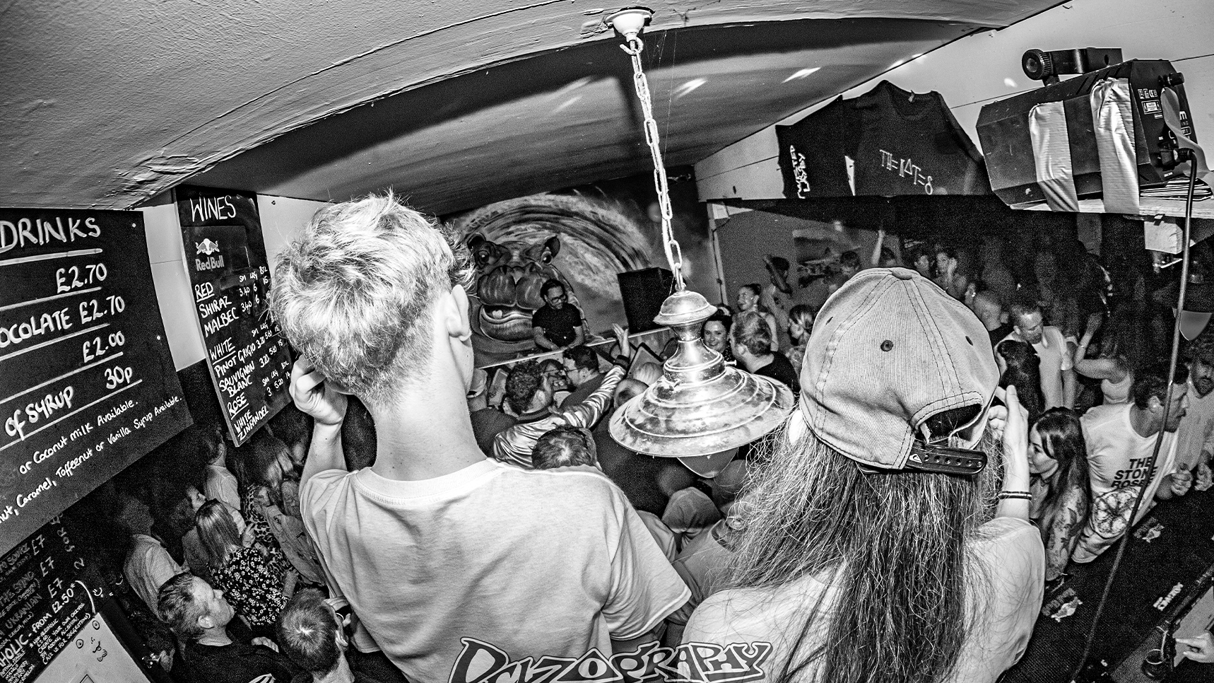 Black-and-white, wide-angle shot of a packed crowd inside the Hippos alternative music venue in Swansea. The audience is gathered in a low-ceiling room facing a DJ performing beneath a large hippo mural, with a chalkboard drinks menu visible on the left.