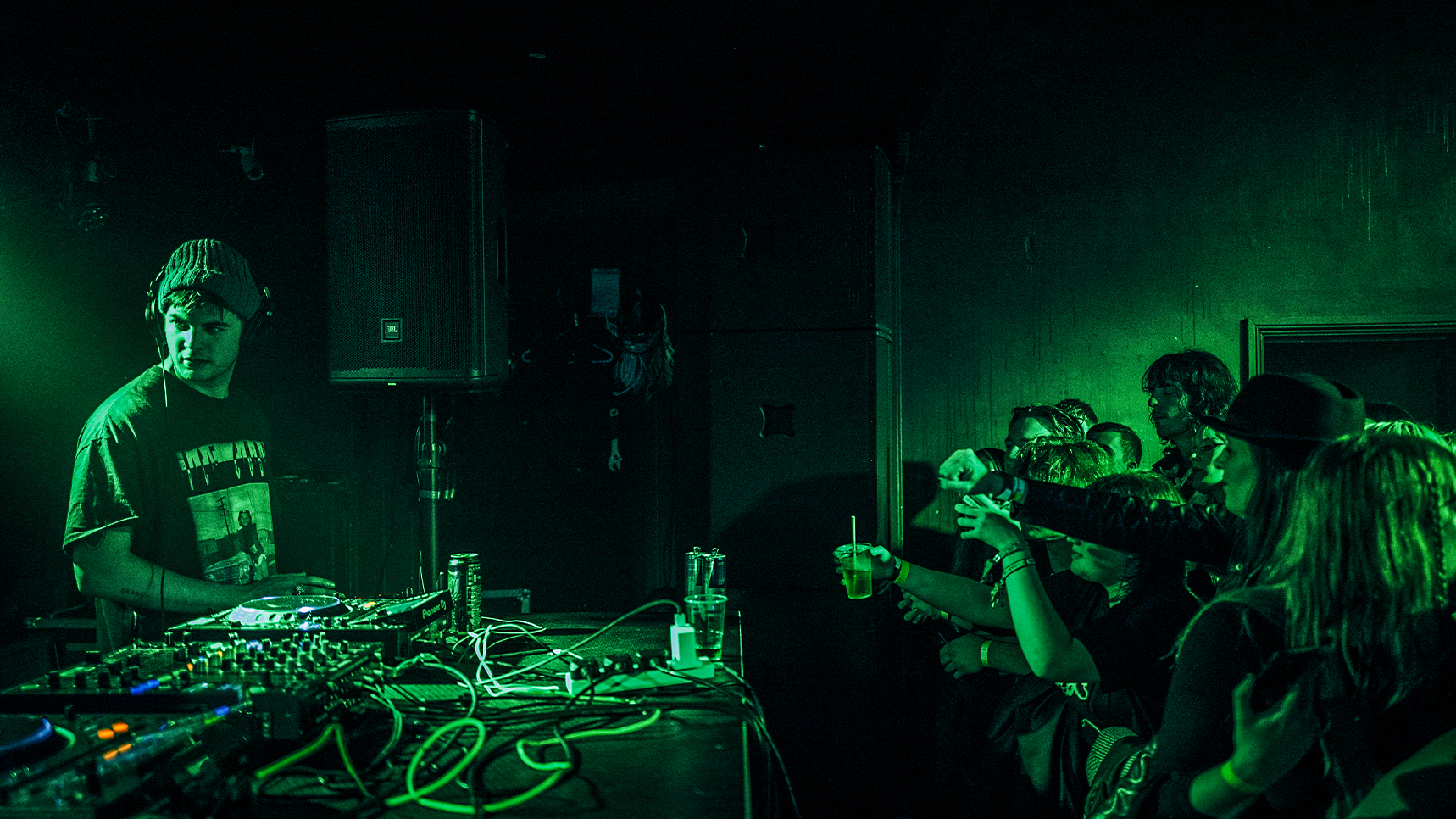 Intimate, green-lit underground DJ set featuring Grian Chatten of Fontaines D.C. at The Bunkhouse in Swansea, looking over a mixing desk at the crow